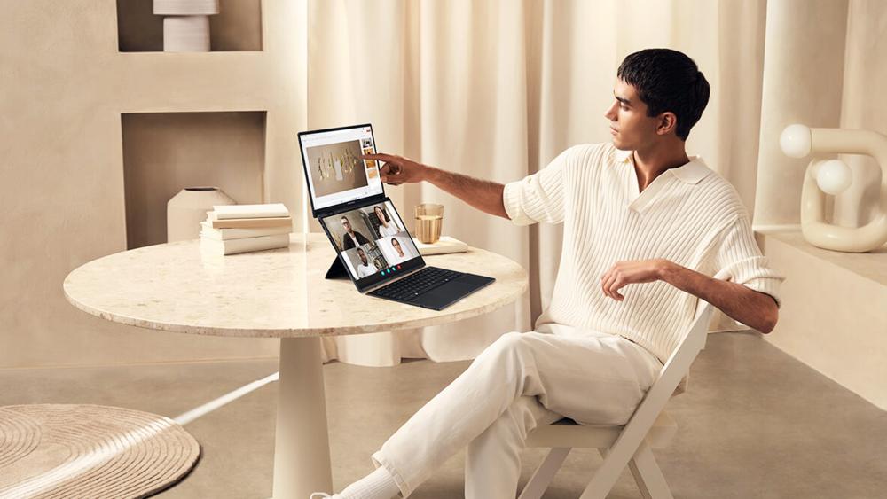Person using an ASUS dual-screen laptop at a table, with a video call on the lower display and content shown on the upper screen.