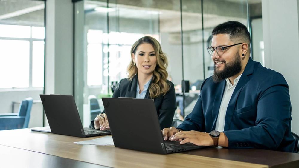 Two business professionals sitting at a meeting table in a modern office, working on laptops.