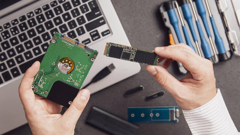 Hands holding HDD and NVMe SSD showing storage upgrade comparison next to laptop and tools for PC performance improvement
