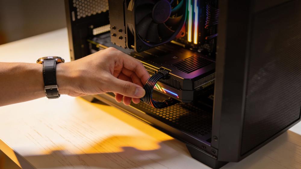 Hand connecting a PCIe power cable to a graphics card inside a desktop PC case with visible CPU air cooler and RGB lighting, illustrating hardware troubleshooting or installation.