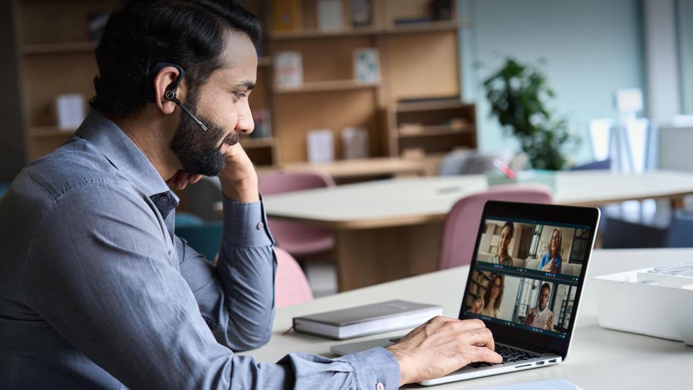 Professional wearing a headset attending a video conference on a laptop in a modern office setting.
