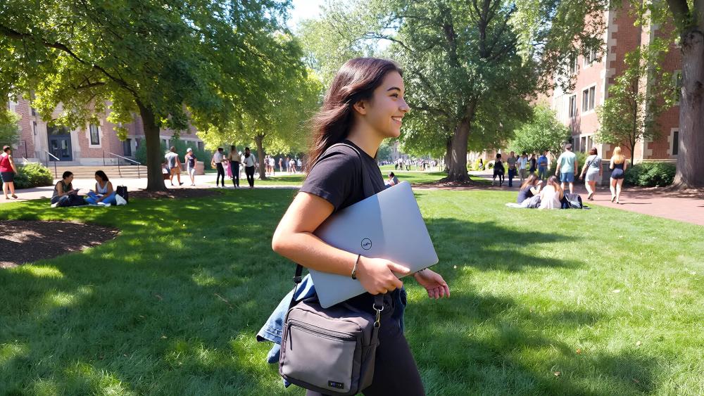 Student walking across a university campus holding a Dell laptop, with trees, grass and other students in the background.