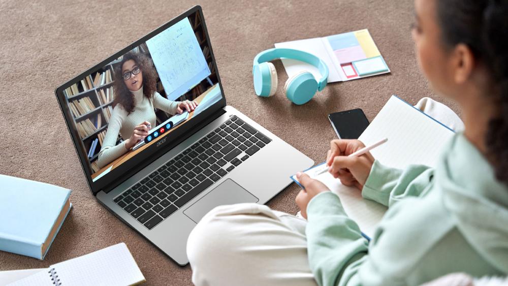 Student taking notes while attending an online class on an Acer laptop, with a teacher visible on a video call screen.