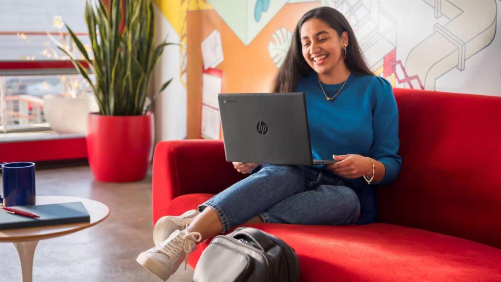 Student sitting on a red sofa using an HP Chromebook laptop in a bright study area with a notebook and backpack nearby.