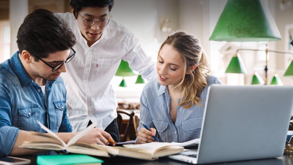 Three students studying together at a table with books and a laptop in a library setting.