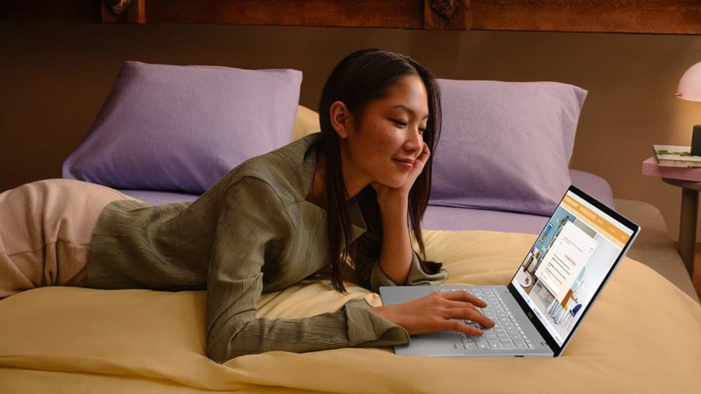 Woman lying on a bed using a slim laptop while browsing or studying online in a cosy bedroom setting.